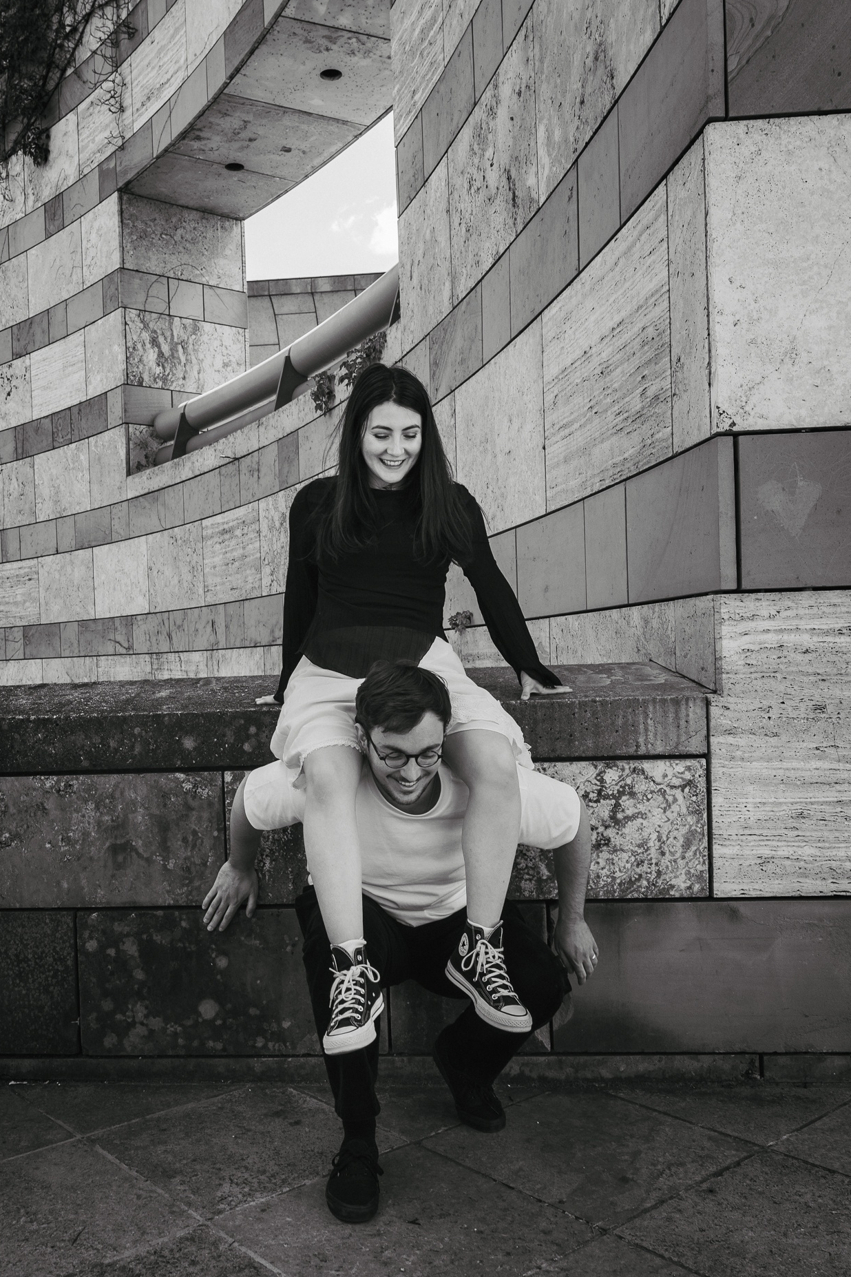 Schwarz-Weiß-Couple-Porträt zweier Personen, eine trägt die andere auf den Schultern vor moderner Architektur