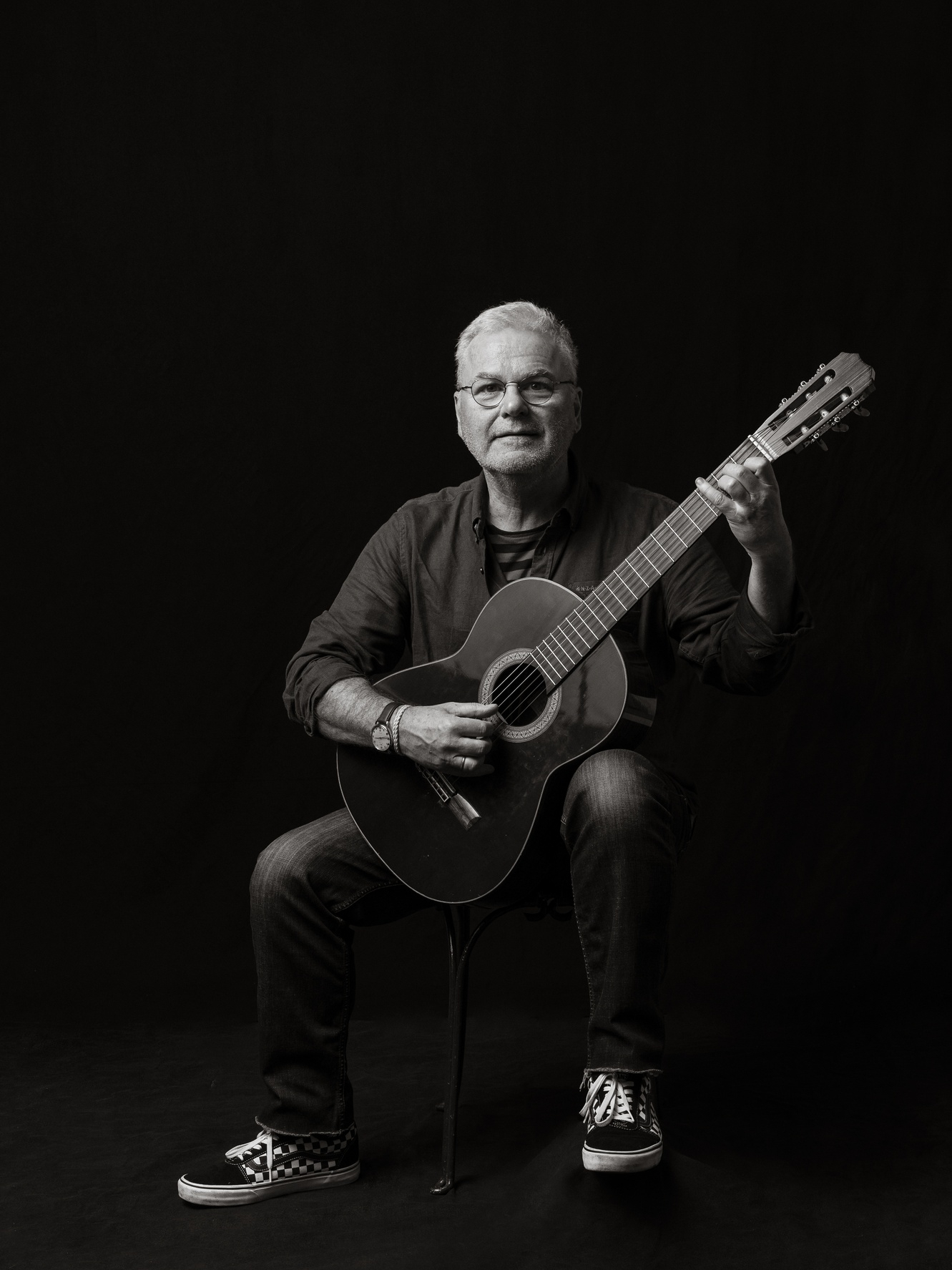 Schwarz-Weiß-Porträt eines Mannes mit klassischer Gitarre in Spielposition vor schwarzem Studiohintergrund