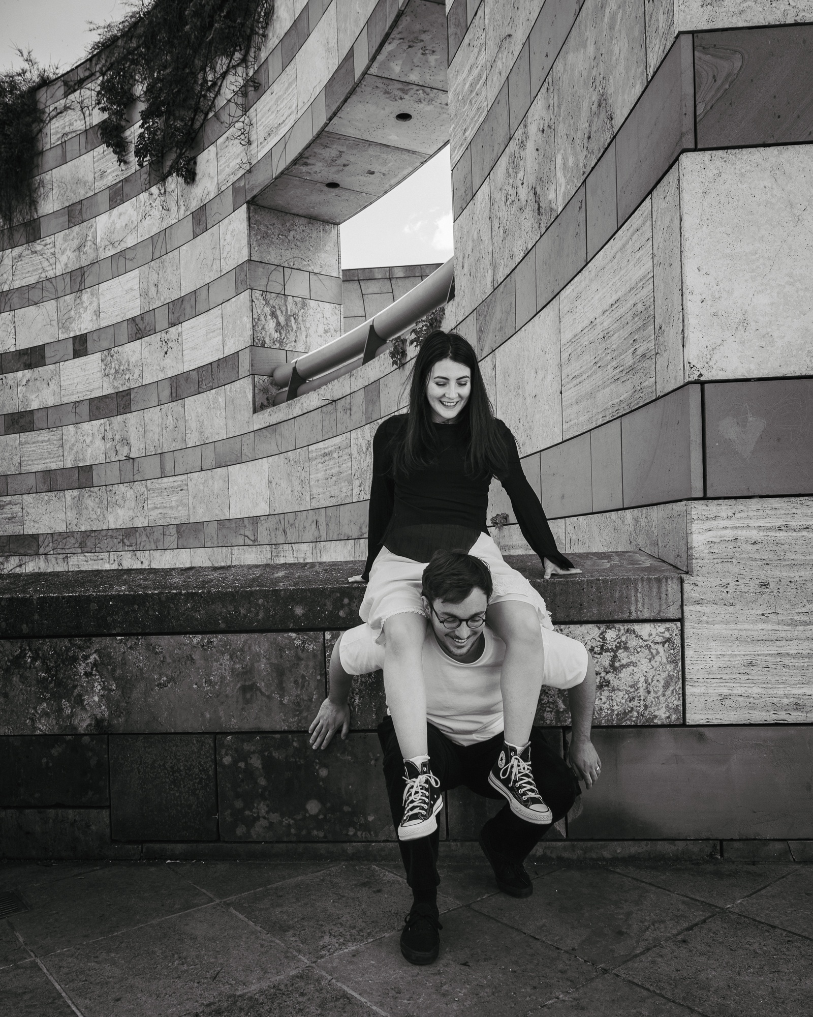 Black and white couple portrait of two people, one sitting on the other's shoulders in front of modern architecture
