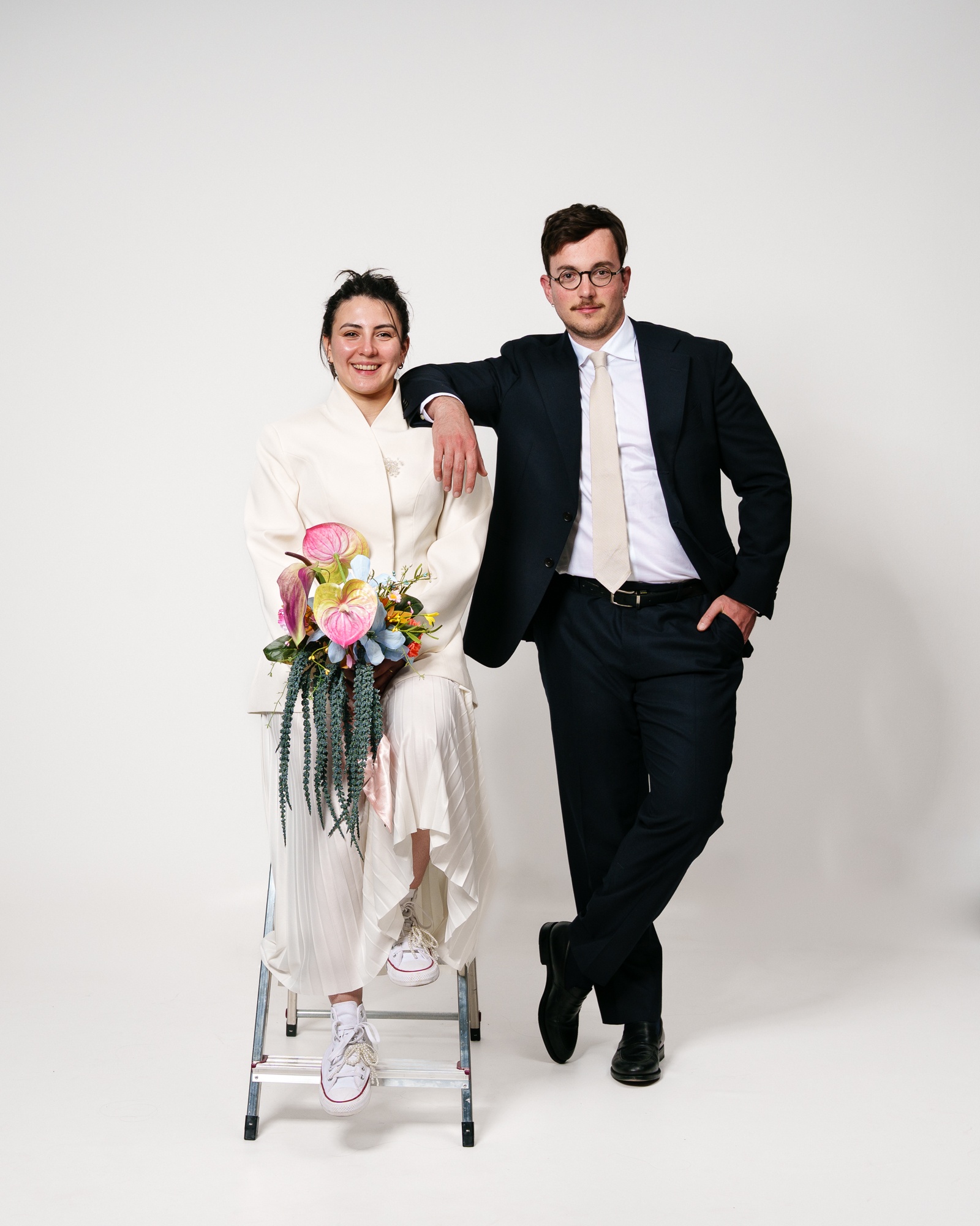 Doppelporträt zweier Personen im Studio, eine sitzt mit einem Blumenstrauß auf einer Leiter, die andere lehnt sich daneben