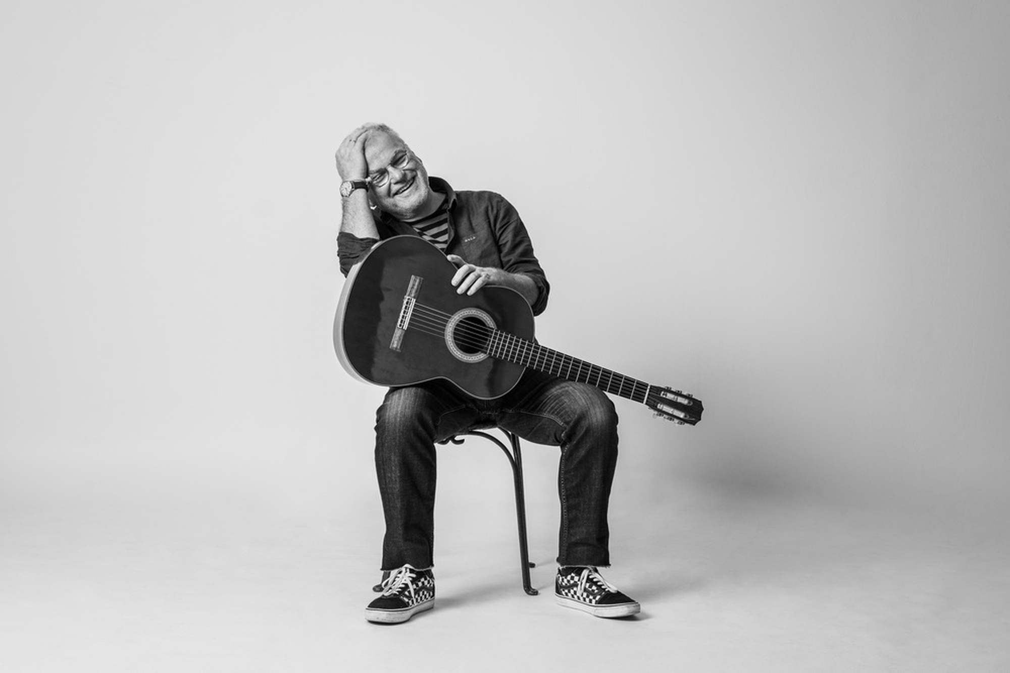Schwarz-Weiß-Porträt eines lächelnden Mannes mit Akustikgitarre auf einem Stuhl vor hellem Studiohintergrund.