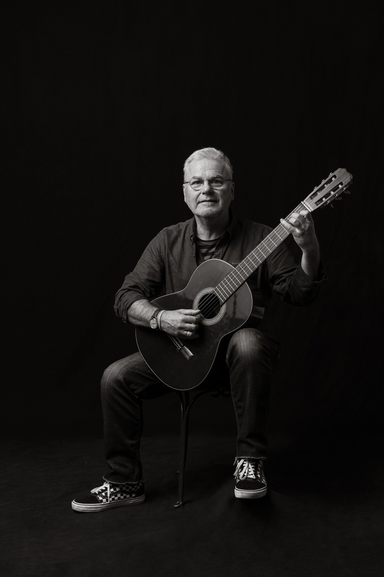 Black and white portrait of a man seated with a classical guitar in playing position against a black studio background