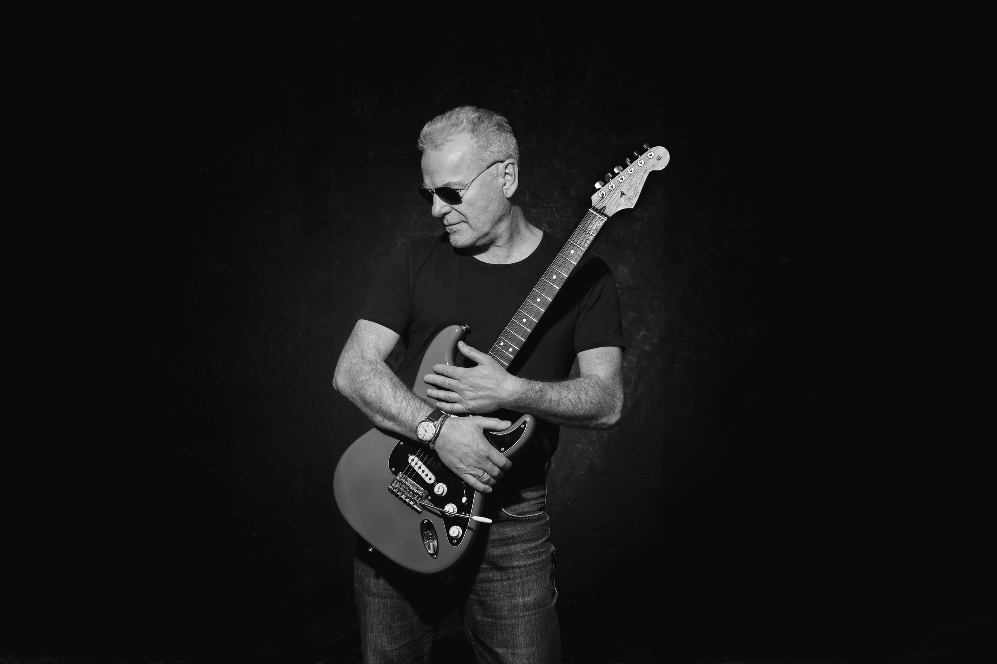 Black and white portrait of a man holding an electric guitar and wearing sunglasses against a black studio background
